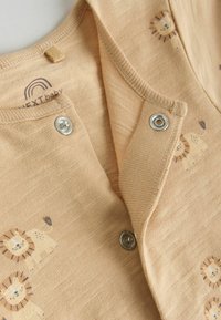 Bež kombinezon za bebe s tiskom lava i metalnim kopčama djelomično otvoren na prednjem izrezu vrata.