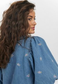 Denimblus med puffärmar, prydd med ömtålig rosa blommönsterbroderi. Tyget har en ljusblå nyans och en mjuk textur.