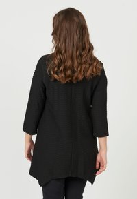 Sort tunika med et tekstureret mønster, trekvartærmer, asymmetrisk søm og en afslappet pasform. Enkel og moderne design.