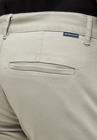 Κοντινή λήψη στην πίσω τσέπη ανοιχτομπεζ παντελονιού με μικρή μαύρη ετικέτα μάρκας Tom Tailor πάνω από τη ραφή της τσέπης.