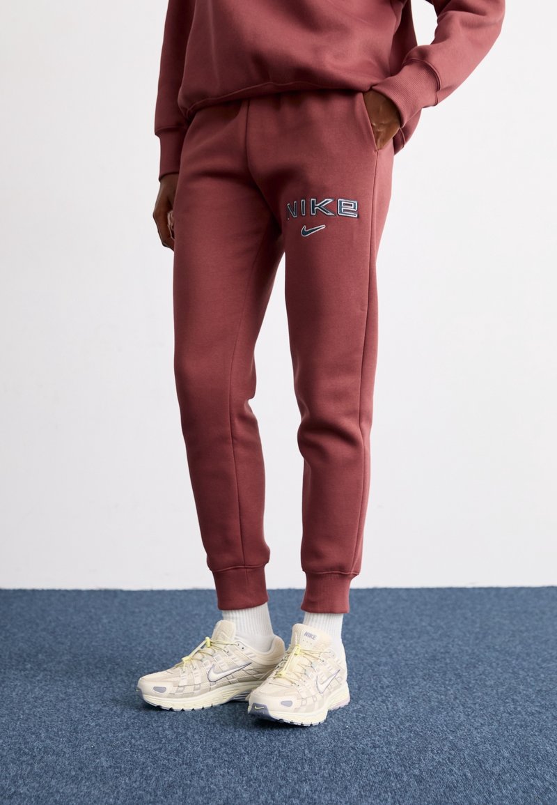 Pantalons de survêtement Nike bordeaux avec des poignets élastiques, portant un logo brodé en bleu clair. Associés à des baskets claires et des chaussettes blanches mi-mollet.