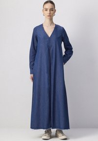 Robe en denim à manches longues, décolleté en V et silhouette droite. Comprend un ourlet effiloché et une fermeture éclair à l'avant, associée à des baskets beiges.