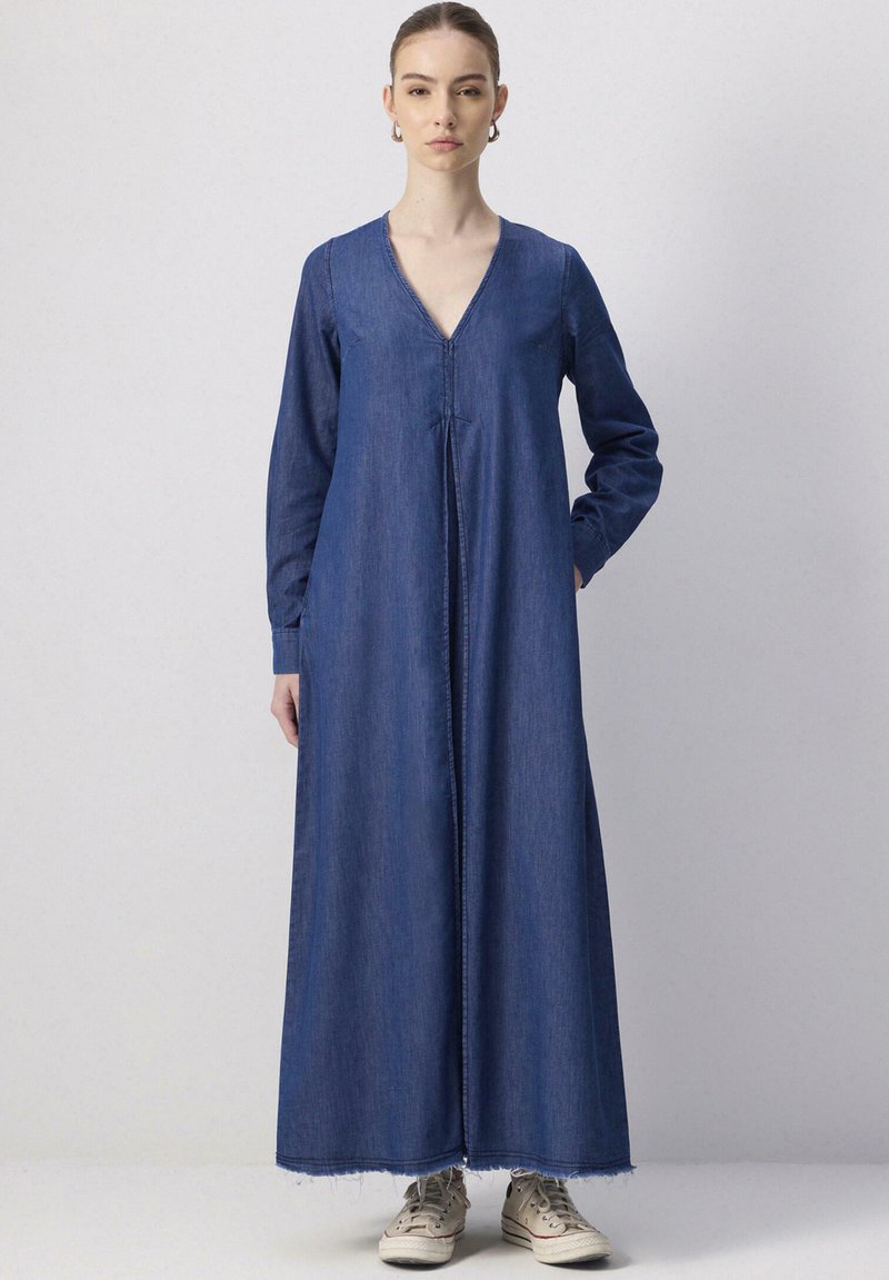 Robe en denim à manches longues, décolleté en V et silhouette droite. Comprend un ourlet effiloché et une fermeture éclair à l'avant, associée à des baskets beiges.