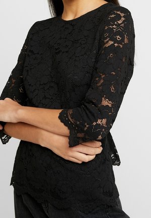 Femme portant un chemisier noir en dentelle florale avec manches transparentes trois-quarts et ourlet festonné, bras croisés.