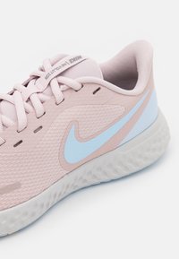 Baskets Nike rose clair avec un dessus texturé, un swoosh bleu et une semelle en caoutchouc ondulée gris clair. Présente un système de laçage et un col rembourré.
