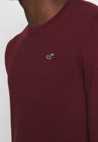 Burgundy tröja med rundad halsringning och ribbade ärmslut, med en liten svart broderad logotyp på bröstet. Mjuk stickad textur.