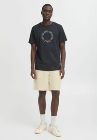 T-shirt en coton bleu marine pour homme avec un texte graphique circulaire gris, associé à un short beige clair et des baskets blanches avec des accents gris.