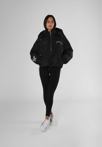Zwarte gewatteerde jas met capuchon, ritssluiting aan de voorkant, logodetails op de borst en linkermouw, gecombineerd met zwarte leggings en witte sneakers.