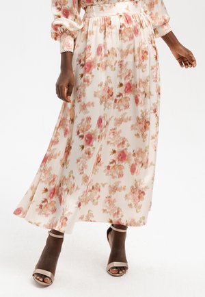 Kobieta w białej, kwiatowej spódnicy midi z różowymi i brzoskwiniowymi kwiatami, w połączeniu z beżowymi sandałami na obcasie z odkrytymi palcami i paskiem wokół kostki.