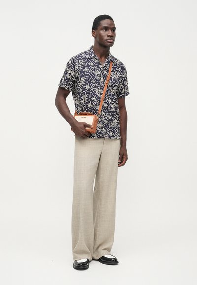 Námornícka modrá košeľa s krátkym rukávom s bielym listovým vzorom, béžové široké nohavice a malá hnedá kabelka cez rameno s remienkom. Čierne topánky s bielymi akcentmi.