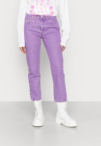 Lila denimjeans med hög midja och rak ben, tillsammans med vita snörda ankelboots. Bakgrunden är enkel och ljusfärgad.