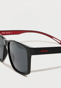 Črne sončne očala z rdečimi poudarki, kvadratna oblika, sivi leč, vsebujejo HUGO logo na templetu. Eleganten plastičen material, gladka tekstura.