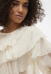 Blouse crème clair avec des volants superposés le long des manches et du col, fabriquée dans un tissu doux et texturé avec un design fluide.