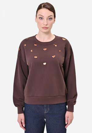Donna che indossa una felpa marrone con paillettes lucide color rame sparse e jeans blu, in piedi davanti a uno sfondo chiaro uniforme.