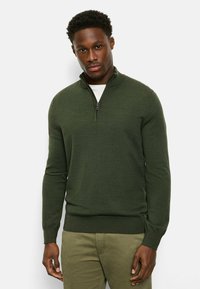 Green quarter-zip sweater made of textured knit fabric, featuring a high collar and ribbed cuffs, worn over a white shirt.