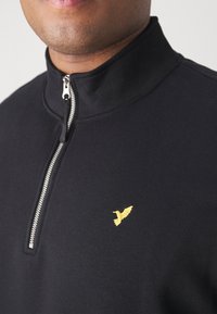 Felpa nera con zip realizzata in materiale morbido. Ha un colletto alto e un logo di un uccello ricamato oro sul petto. La zip è di colore argento.