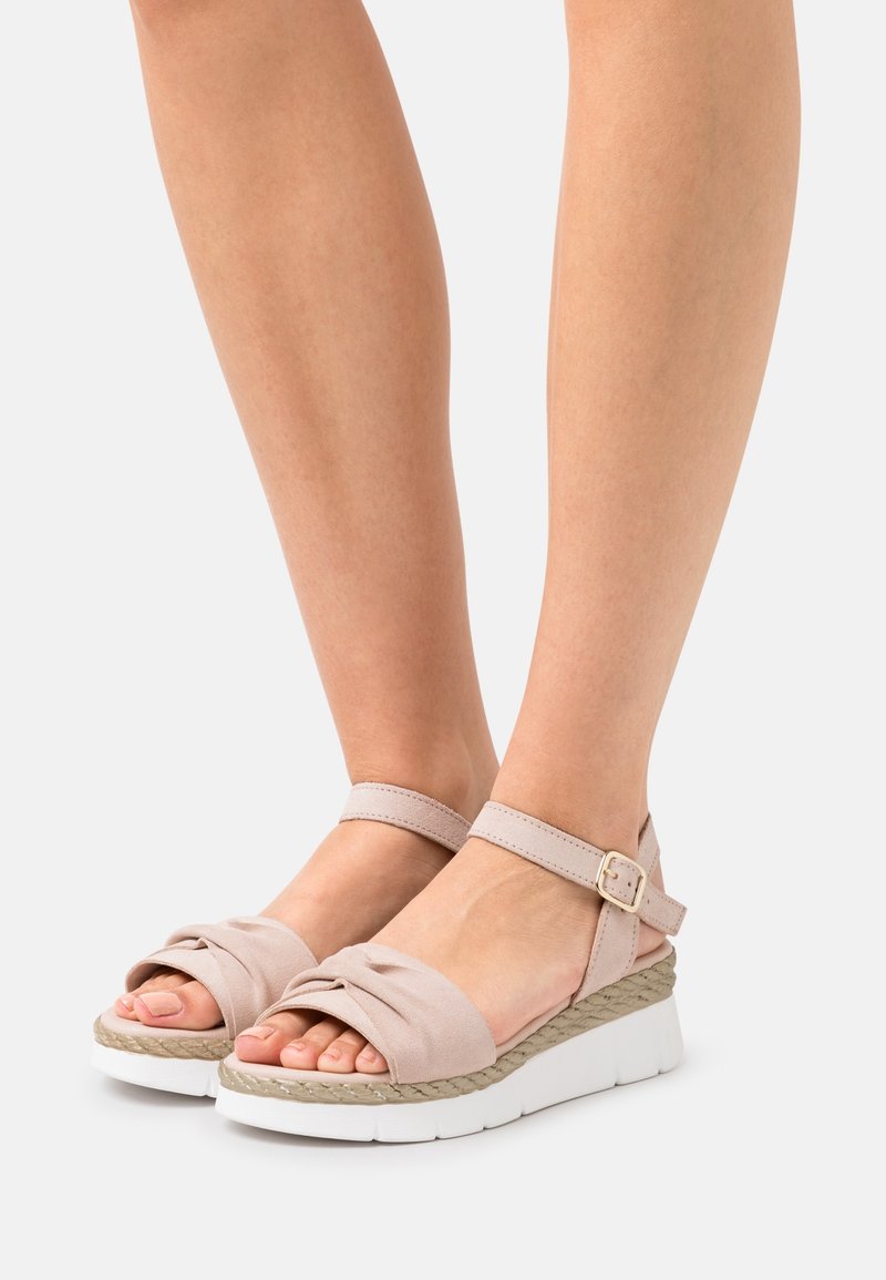 Pieds portant des sandales beige à bout ouvert avec des brides à la cheville, une semelle intermédiaire texturée et des semelles plateformes blanches sur un fond clair.