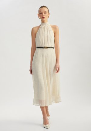 Vestito halter di crema con pois neri, scollatura arricciata e una cintura nera aderente con accessori dorati. Lunghezza a metà polpaccio e texture trasparente.