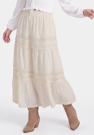 Vrouw met een lange crèmekleurige kanten rok met gelaagde borduursels en lichtbruine suède enkellaarsjes, vanaf de taille naar beneden getoond op een witte achtergrond.