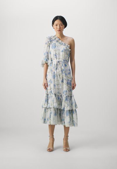 Floral maxi-jurk met een schouderdesign, ruffles en een gelaagde zoom. De stof heeft blauwe bloemen op een lichte achtergrond. G gedragen met neutrale hakken.