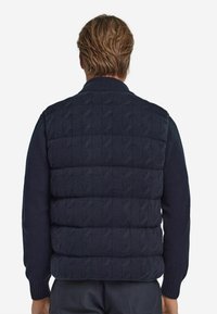 Granatowa dzianinowa kamizelka z wzorem warkocza na górnej części i pikowaną dolną połową; posiada prążkowany kołnierz i otwory na ramiona.