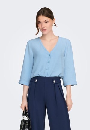 Femme portant un chemisier boutonné bleu clair à col en V et un pantalon taille haute bleu marine, tenant un sac à main noir sur un fond uni.