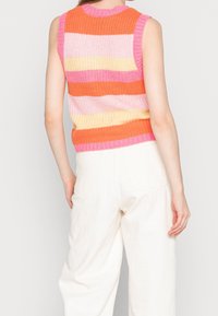 Ærmeløs strikket top med vandrette striber i orange, pink og gul, båret med højtaljede hvide bukser, vist fra ryggen.