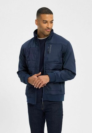 Homme portant une veste matelassée bleu marine et un jean foncé, debout les mains jointes, regardant vers sa gauche sur un fond uni.