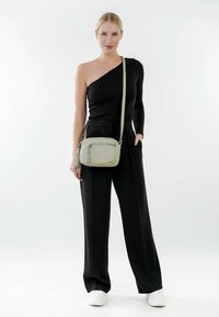 Borsa a tracolla verde chiaro con superficie liscia, tasca con zip e tracolla regolabile. Il modello indossa un top nero a spalla unica e pantaloni a gamba larga.