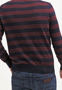 Mann trägt einen bordeauxroten und dunkelblauen horizontal gestreiften Pullover mit gesprenkeltem Muster und dunkelblaue Jeans, von hinten gezeigt.