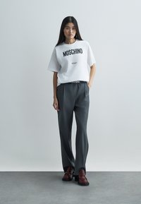 Bel beli kratkotrajni majica s črnim potiskom "Moschino Milano", združena s temno sivimi hlačami in bordo rdečimi natikači na teksturiranem tleh.