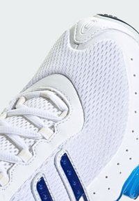 Bijela atletskih tenisica s mrežastim i kožnim materijalima, složenim sustavom vezanja, plavim detaljima sa strane i teksturiranom potplatom.
