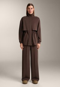 Rjava krajena jakna z visokim ovratnikom in vrvico v pasu, združena s širokimi hlačami iz ujemajočega tkanine. Gladek tekstura, minimalni detajli.