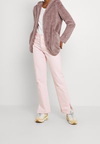 Felpa in pelliccia sintetica di un accogliente colore viola chiaro con colletto a scialle, abbinata a pantaloni a zampa d'elefante di un tenue rosa pallido. Le scarpe sono delle sneakers sportive beige e argento.