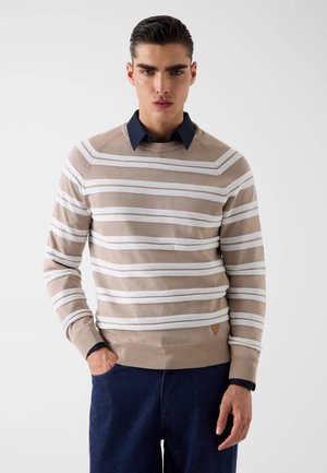 Joven con cabello oscuro peinado hacia atrás, que lleva un suéter a rayas beige y blanco sobre una camisa azul marino con cuello, de pie con una mano en el bolsillo.