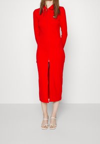 Robe rouge zippée avec col, longueur mi-mollet, manches longues, tissu lisse, présentant une fente à l'avant et un petit logo sur la poitrine.