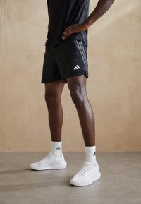 Svarta atletiska shorts med tre vita ränder på sidan, kombinerat med vita sneakers och ribbade vita strumpor med en logotyp.