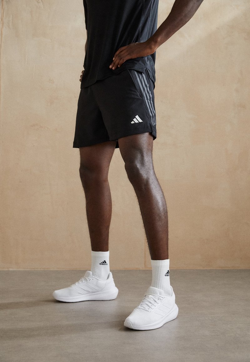 Svarta atletiska shorts med tre vita ränder på sidan, kombinerat med vita sneakers och ribbade vita strumpor med en logotyp.