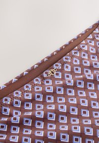 Gros plan sur un tissu marron avec un motif carré bleu et blanc, présentant un petit logo métallique circulaire doré près du bord cousu.