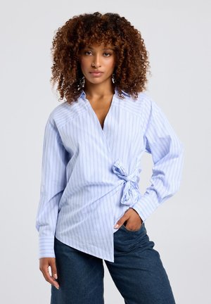 Vrouw met krullend haar die een lichtblauw gestreept overslagshirt draagt, gestrikt in de taille, en donkerblauwe jeans, poserend met één hand in de zak.