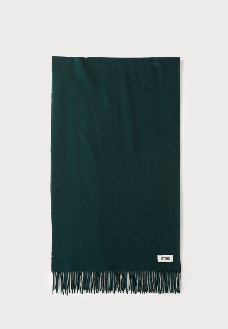Écharpe vert foncé en tissu doux, dotée de longues franges aux extrémités et d'une petite étiquette blanche avec du texte noir sur un côté.