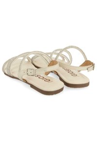 Beige sandaler med krydsede remme, der har sølv accenter. Flad sål med en spændet ankelrem og en polstret sål.