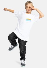 Vit t-shirt med en mångfärgad "DCShoes Company" logotyp, matchad med svarta jeans och grå sneakers med vita snören och teal detaljer.