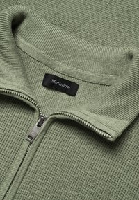 Pull vert à fermeture éclair, tissu texturé, col et ourlet côtelés, avec une fermeture éclair argentée et une étiquette noire portant le logo "Matiniqu".