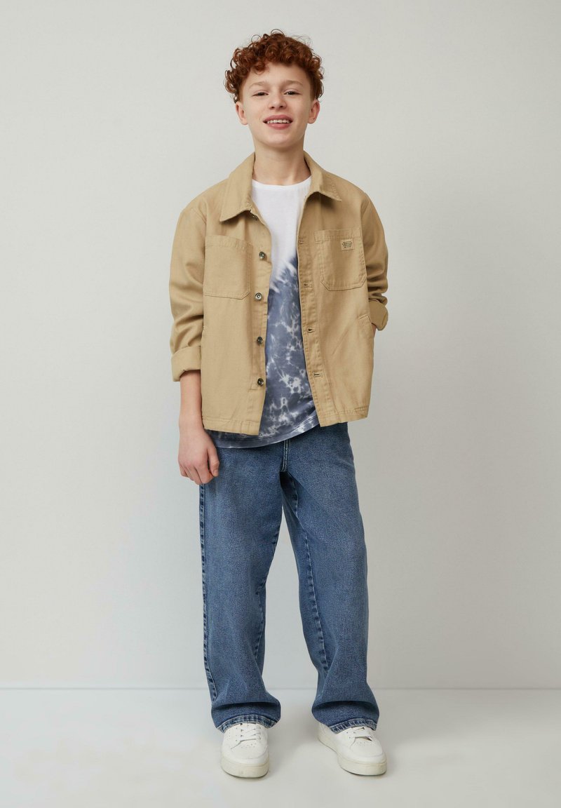 Lichtbeige button-up jasje over een wit en blauw tie-dye shirt, gecombineerd met losse blauwe denim jeans en witte sneakers.