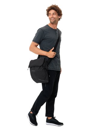 Borsa a tracolla nera con chiusura a pattina, tracolla regolabile e tessuto strutturato. Indossata da una persona in abbigliamento atletico nero e sneakers.
