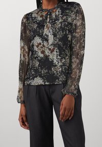 Donna che indossa una blusa nera a fiori con maniche trasparenti e un nodo legato al collo, abbinata a pantaloni grigi scuri.