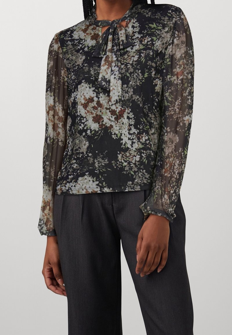 Donna che indossa una blusa nera a fiori con maniche trasparenti e un nodo legato al collo, abbinata a pantaloni grigi scuri.
