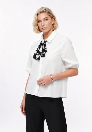 Γυναίκα που φοράει λευκό μπλουζάκι με κοντά μανίκια και μαύρη φλοράλ γραβάτα στον λαιμό, μαύρο παντελόνι, ασημένιο βραχιόλι και κοντά ξανθά μαλλιά, μπροστά σε λευκό φόντο.