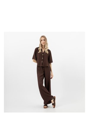 Vrouw met lang golvend blond haar, gekleed in een losse bruine blouse met knopen en bijpassende wijde broek, staand met één been gekruist over het andere.